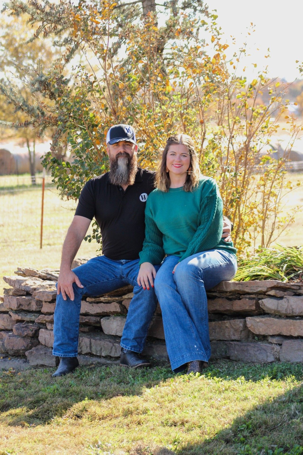 Couple sitting on a stone wall outside; man with beard, blue jeans, woman in green sweater, autumn setting.