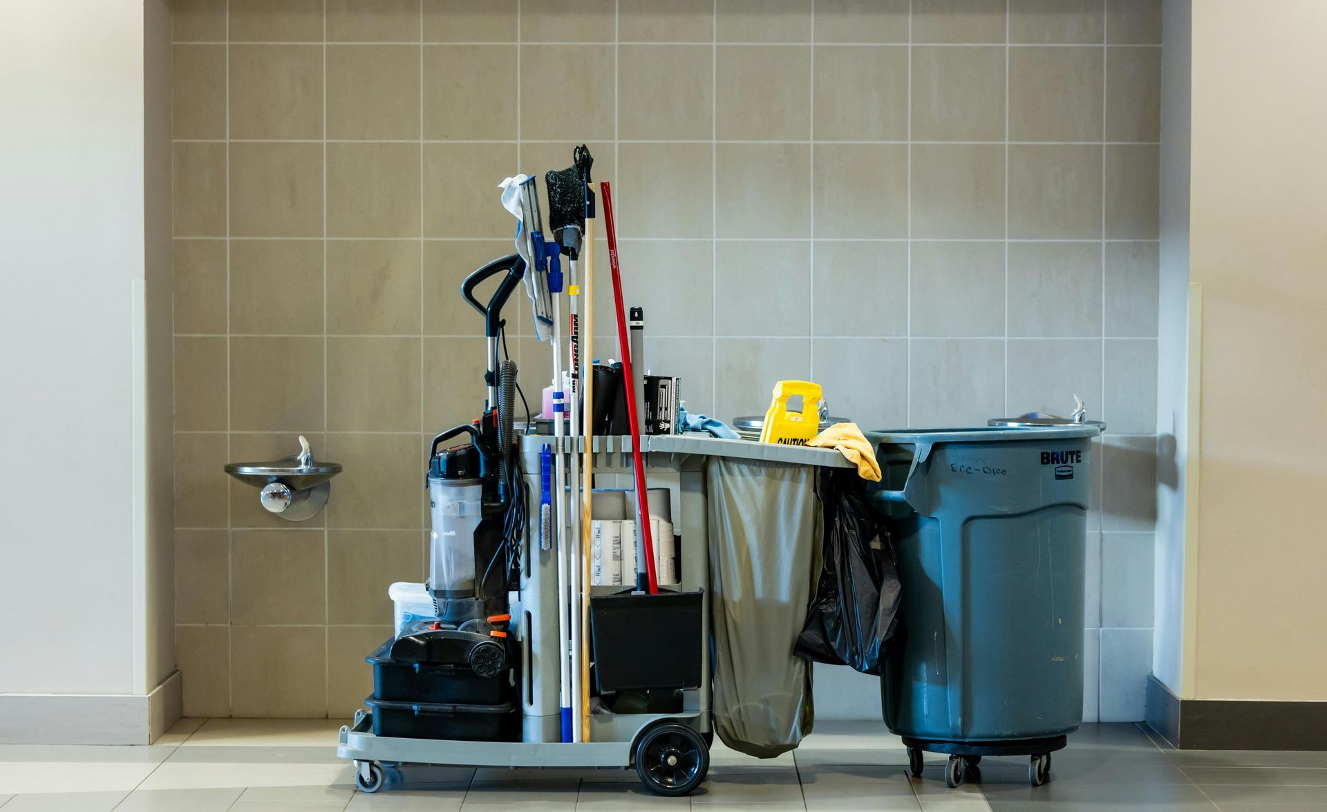 Cleaning cart with various supplies, next to a trash can in a hallway.