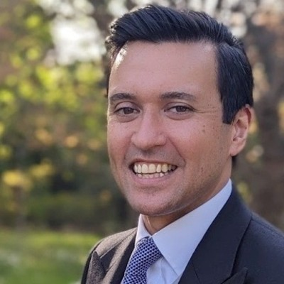 A smiling person in a dark suit, light shirt, and patterned tie, set against an out-of-focus natural green background.
