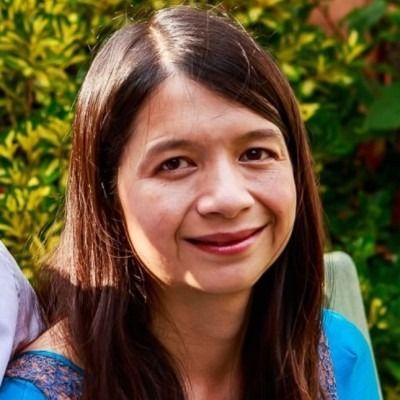 A smiling person with long dark hair wearing a bright blue top against a blurred background of green foliage.