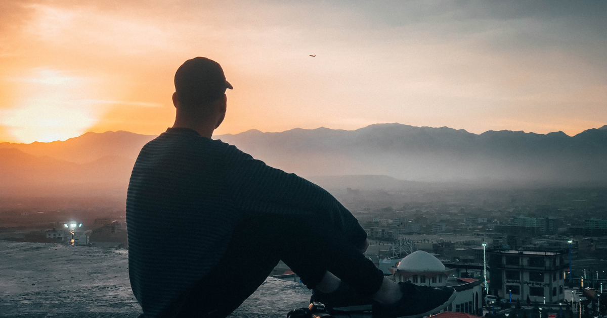 Person in a cap sits atop a building overlooking a city at sunset.