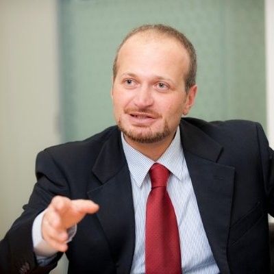 Man in a dark suit and red tie speaking and gesturing in an office setting