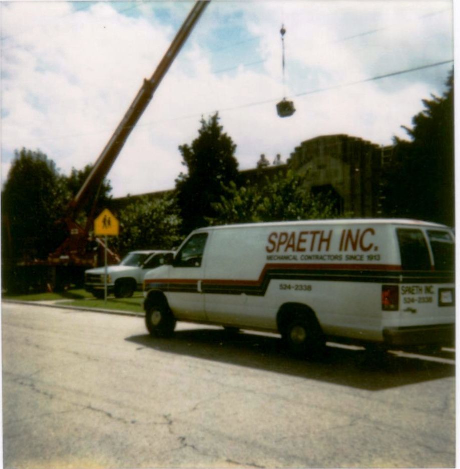 Two gray air conditioning units beside a beige building, set on gravel and concrete, with green grass.