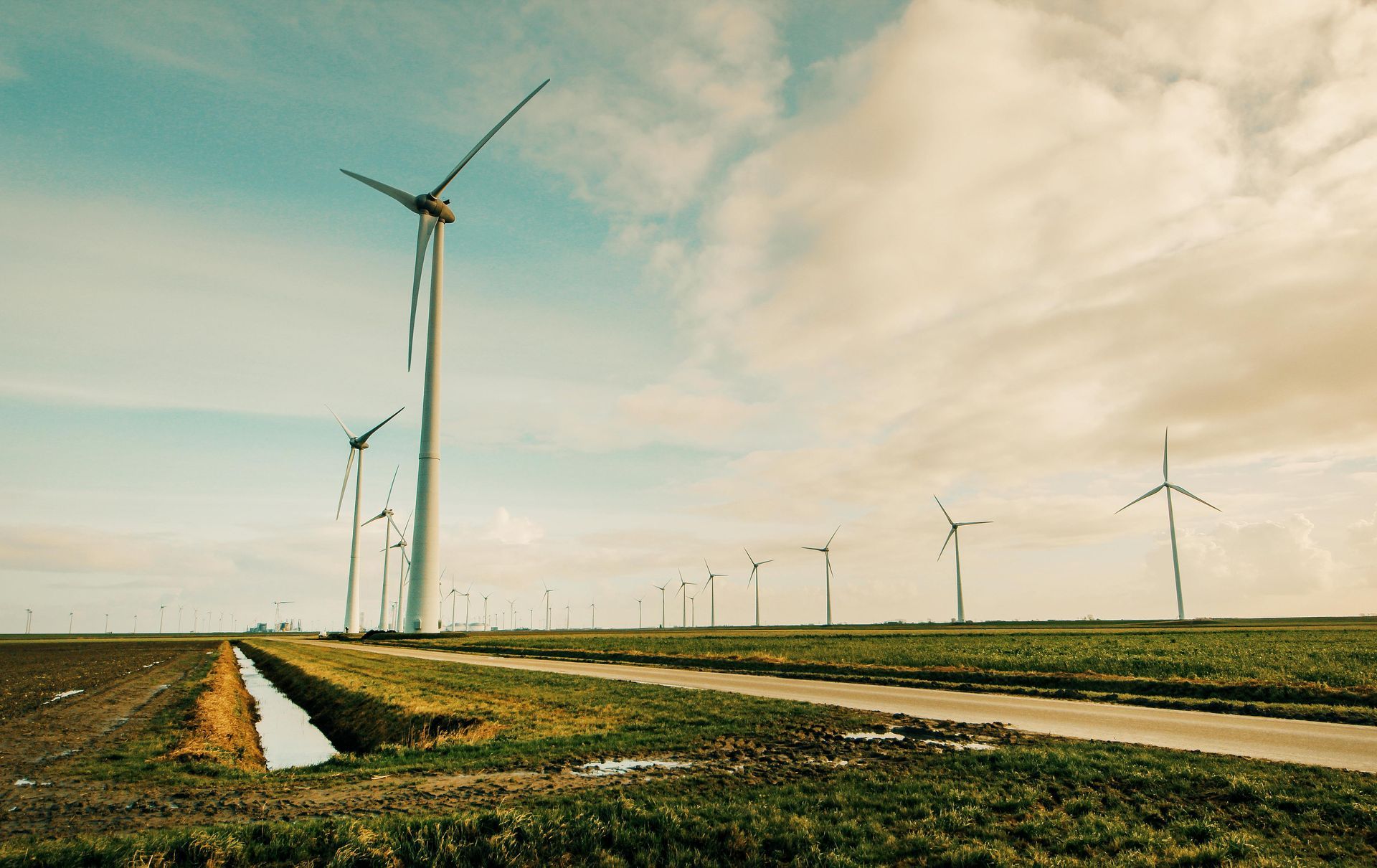 Wind turbines in a rural field under a cloudy sky. A ditch and road run parallel to the turbines.