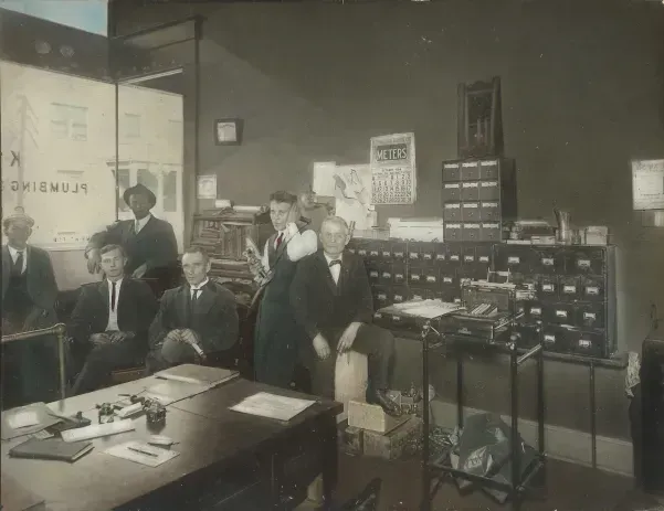 Group of men in suits in an office; men at desk, cabinets, and on the sides; items on shelves.