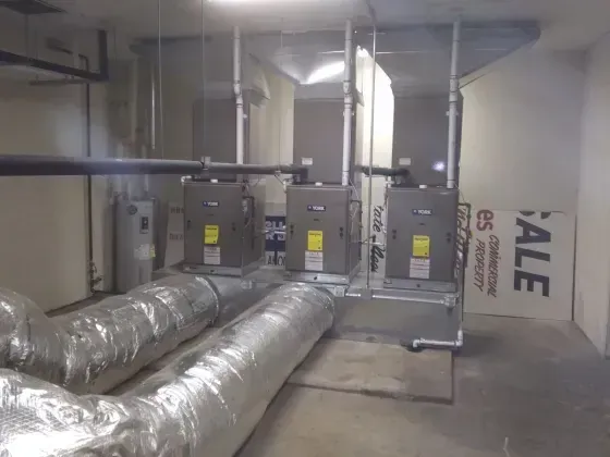 Three HVAC units on a metal rack, with insulated silver ducts and pipes in an industrial space.