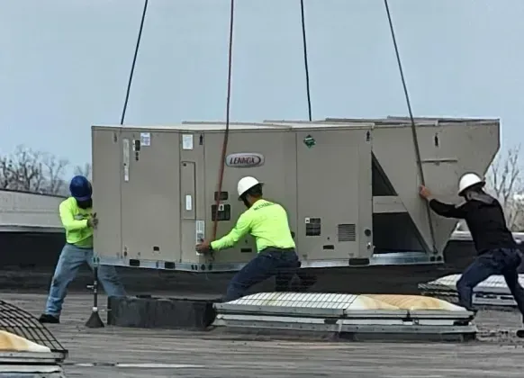 Workers in hard hats and neon shirts lifting HVAC unit onto a rooftop with a crane.