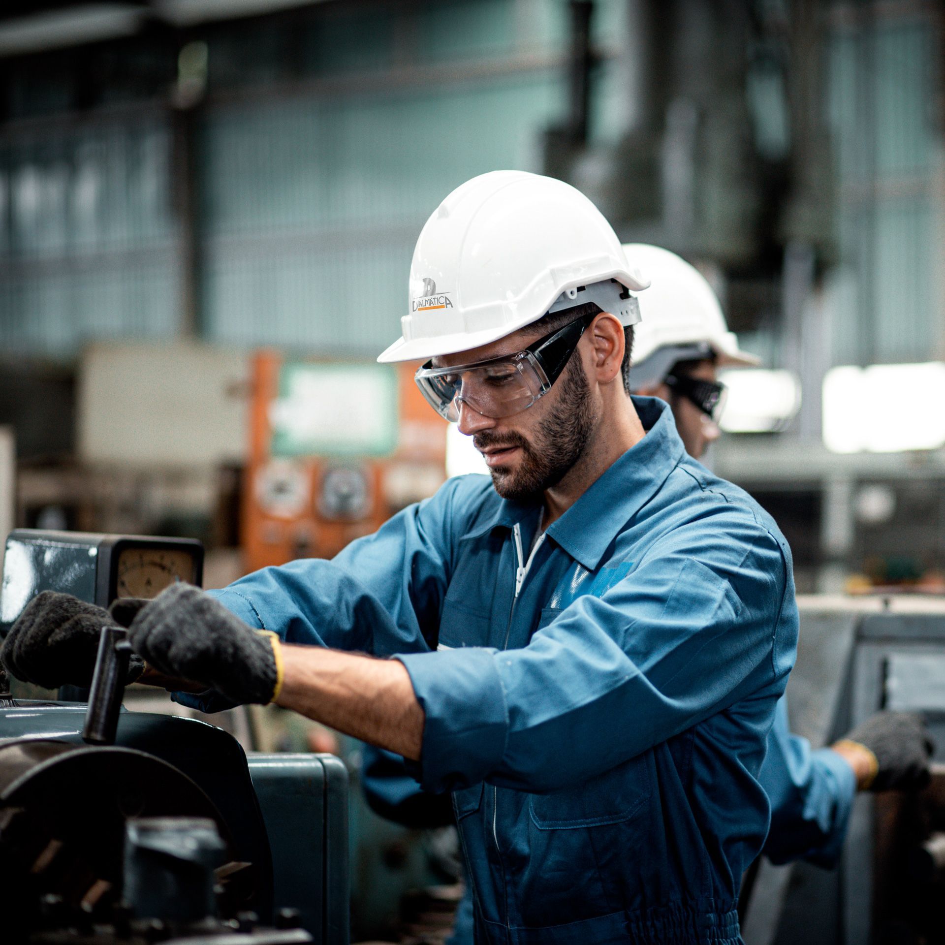 Um homem de macacão azul e capacete opera máquinas em uma fábrica, usando óculos de segurança e luvas.