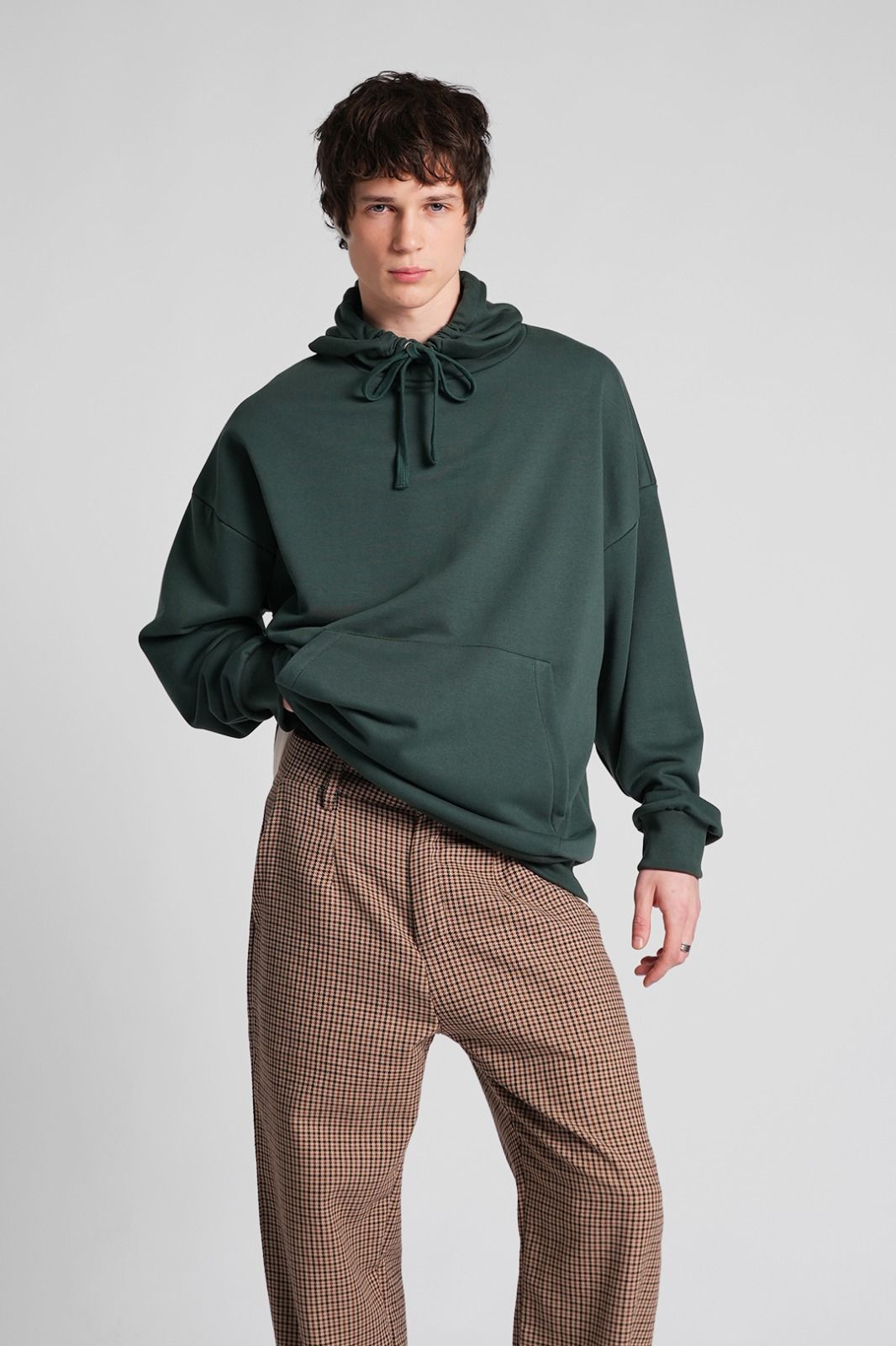 Hombre con sudadera con capucha verde y pantalones a cuadros, de pie sobre un fondo gris.