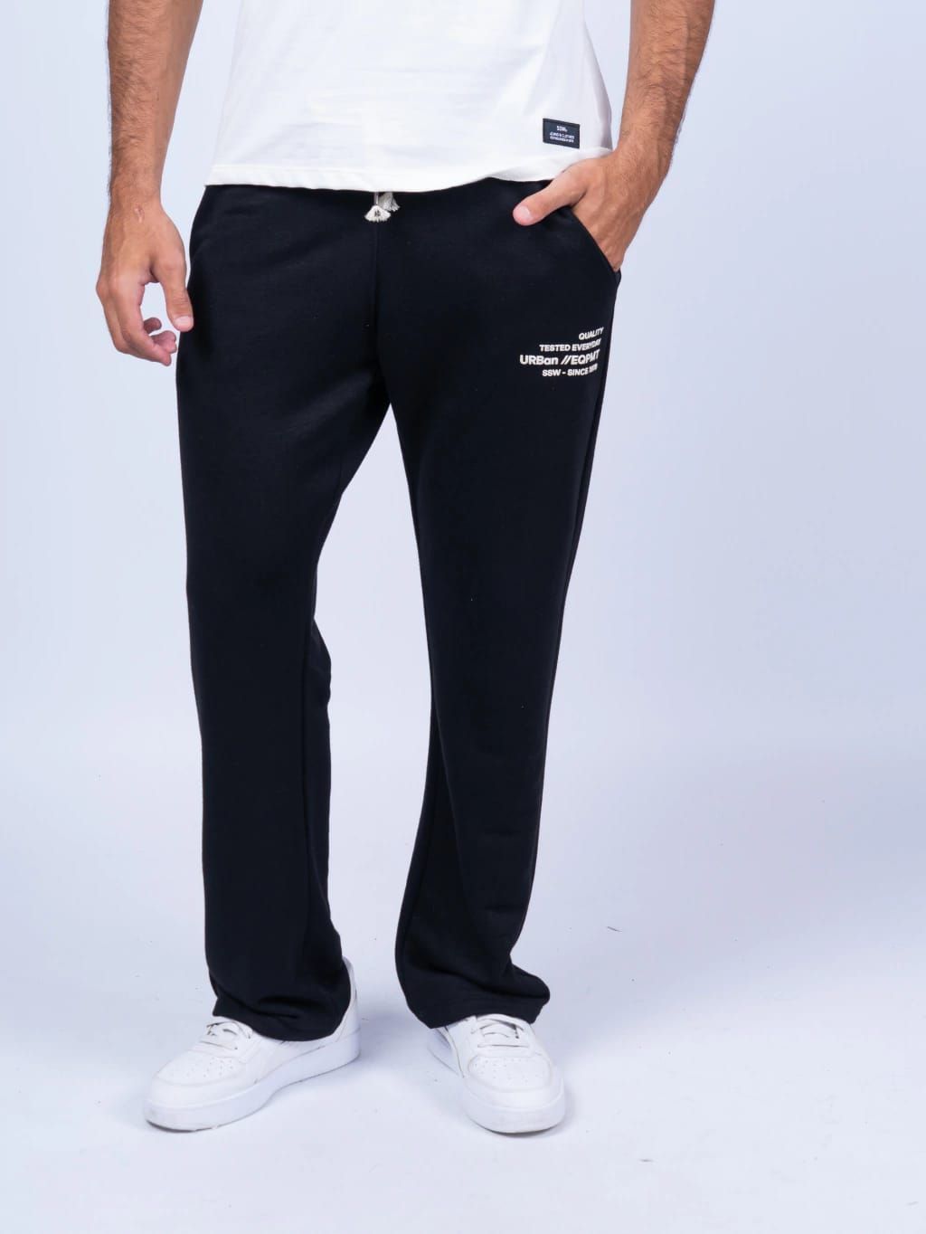 Hombre con pantalones deportivos negros, camiseta blanca, zapatillas blancas y mano en el bolsillo.