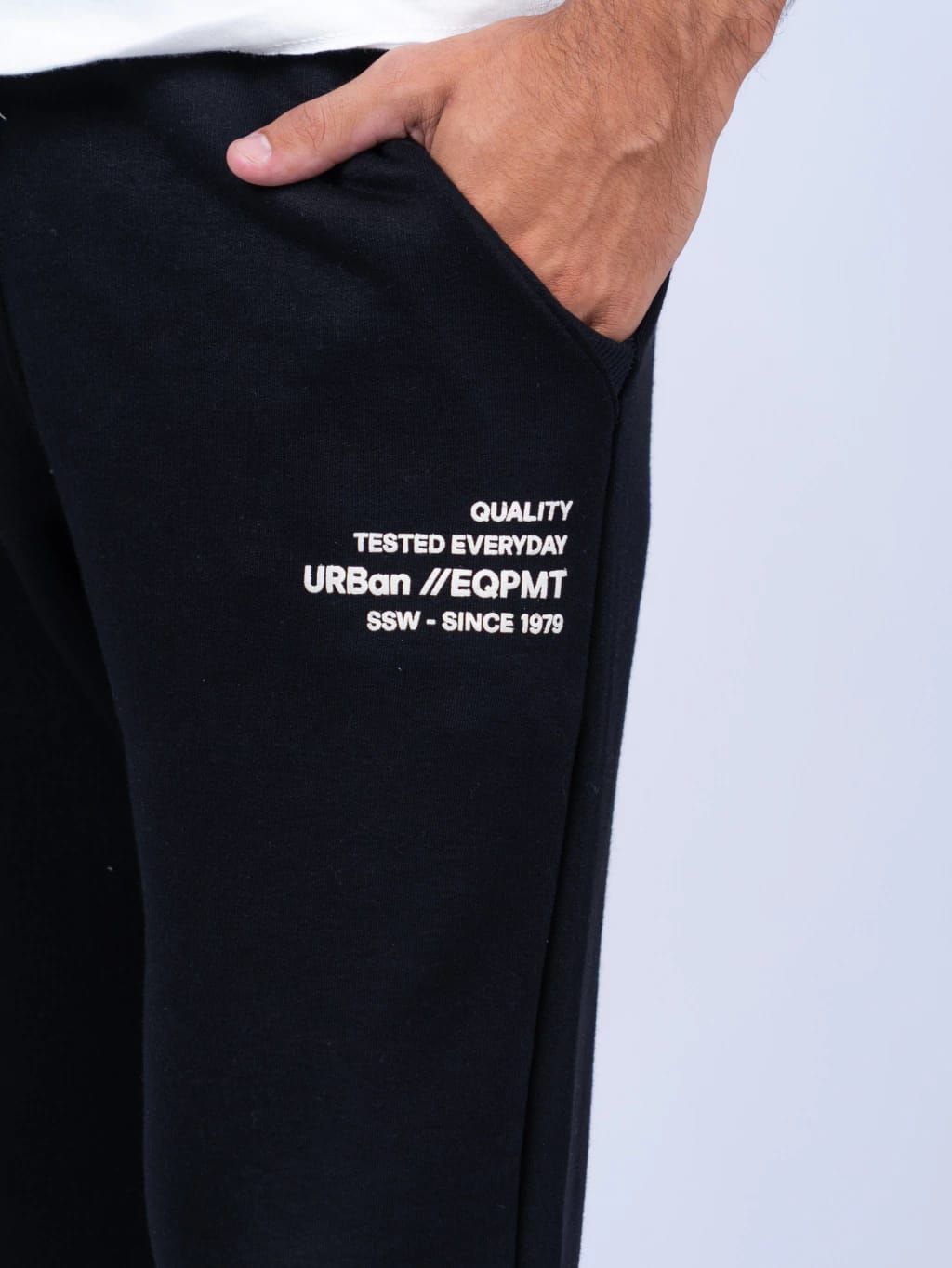 Hombre con pantalones deportivos negros, camiseta blanca, zapatillas blancas y mano en el bolsillo.