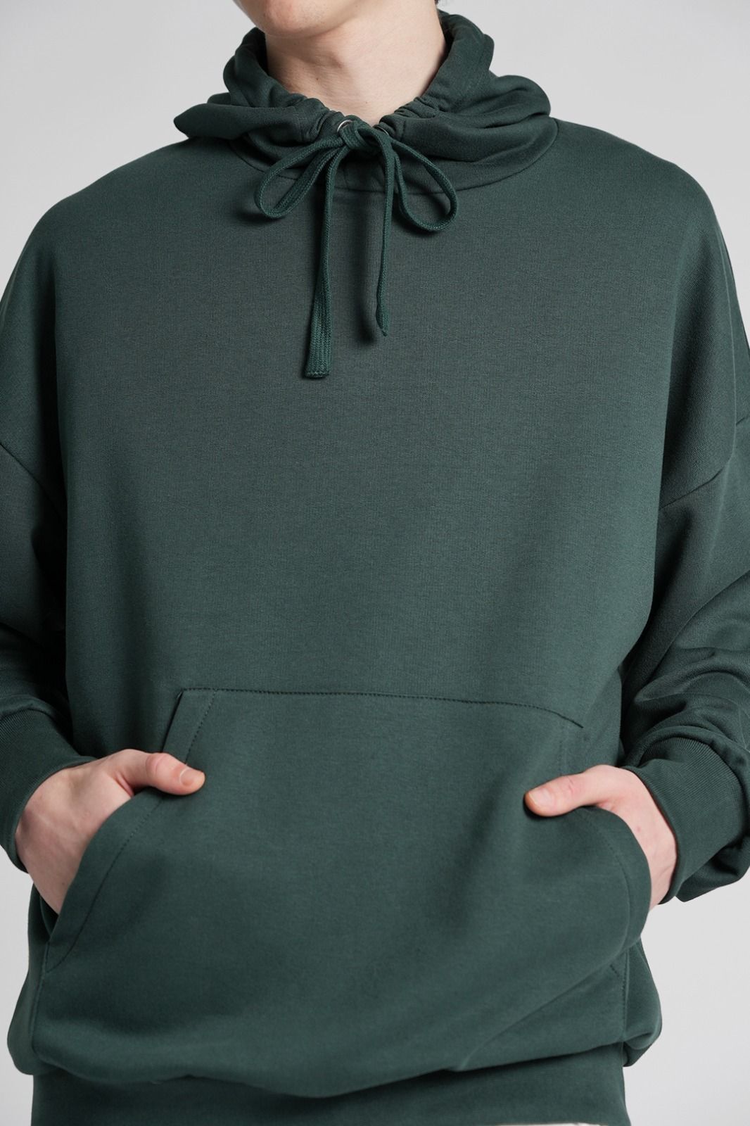 Hombre con sudadera con capucha verde y pantalones a cuadros, de pie sobre un fondo gris.