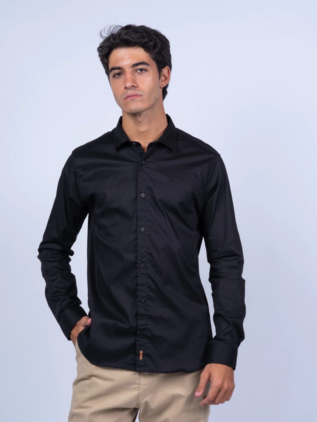 Hombre con camisa negra abotonada y pantalones caqui, mano en el bolsillo, mirando hacia adelante.