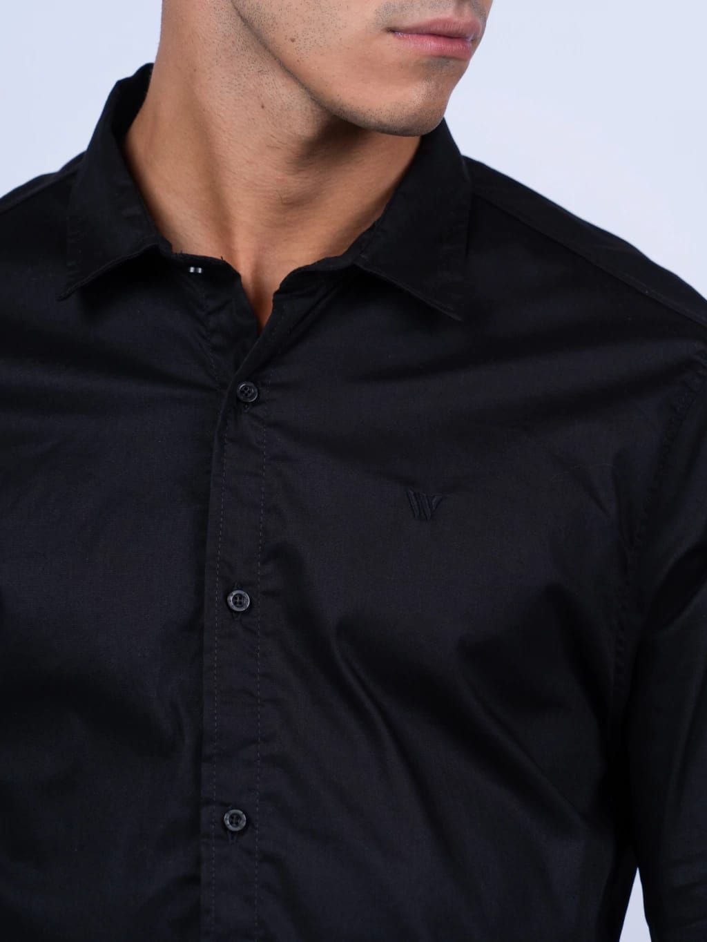 Hombre con camisa negra abotonada y pantalones caqui, mano en el bolsillo, mirando hacia adelante.