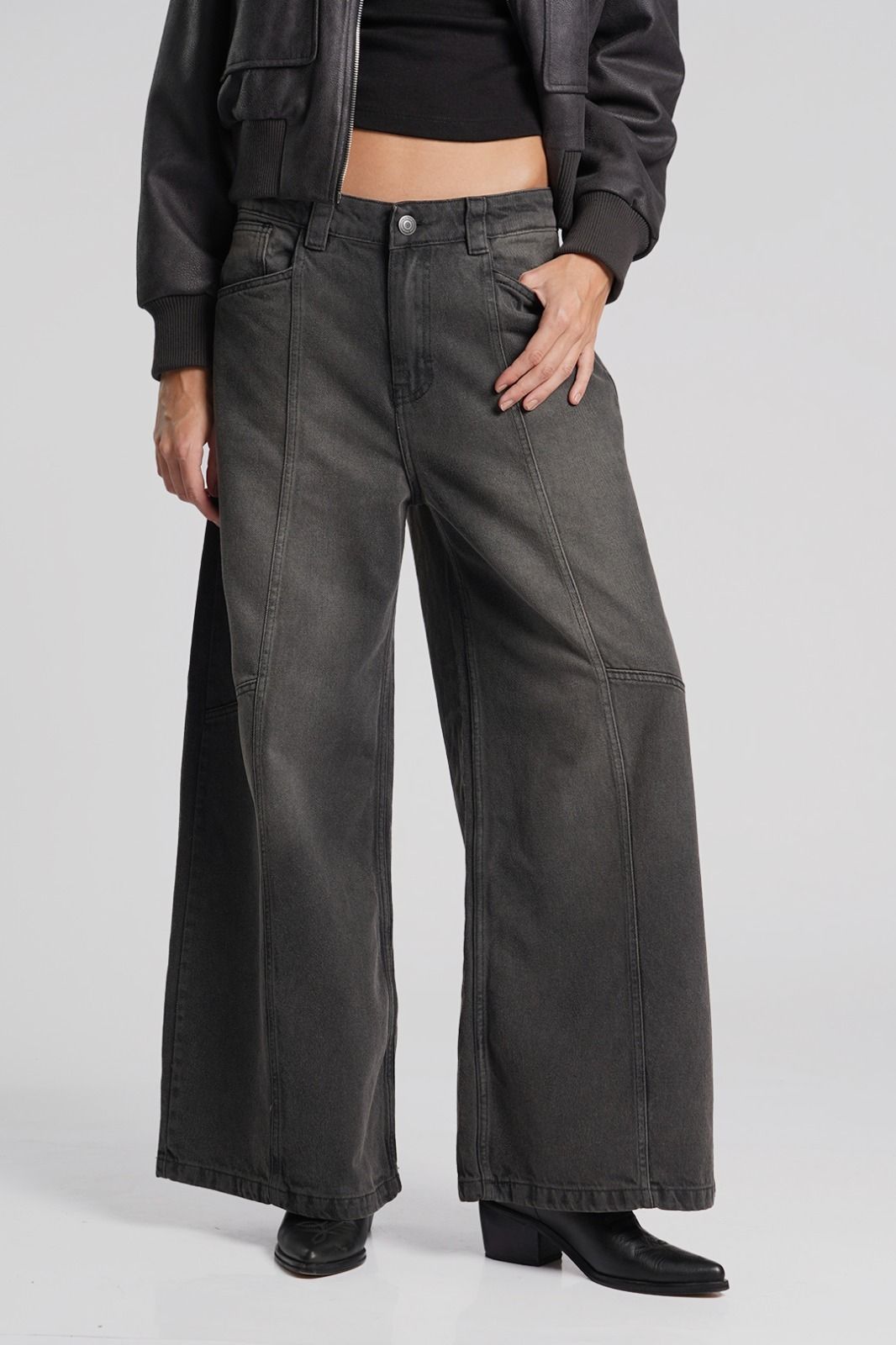 Mujer con chaqueta gris oscuro y jeans de pierna ancha, de pie frente a un fondo blanco.