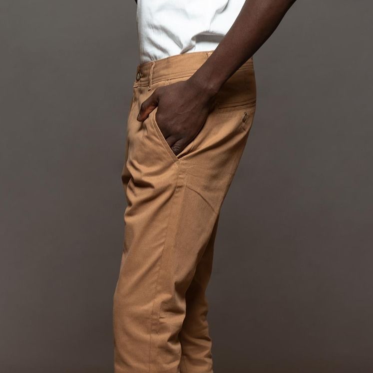 Hombre vestido con pantalones marrones y camisa blanca, de pie sobre un fondo neutro.