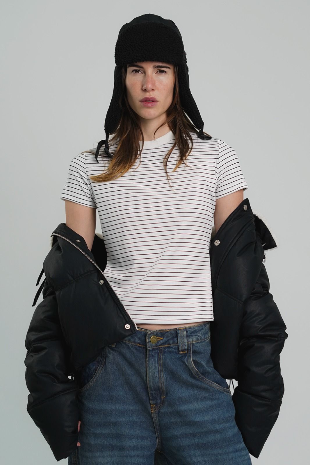 Mujer con sombrero negro, camisa de rayas blancas, jeans azules y chaqueta negra sobre un fondo blanco.