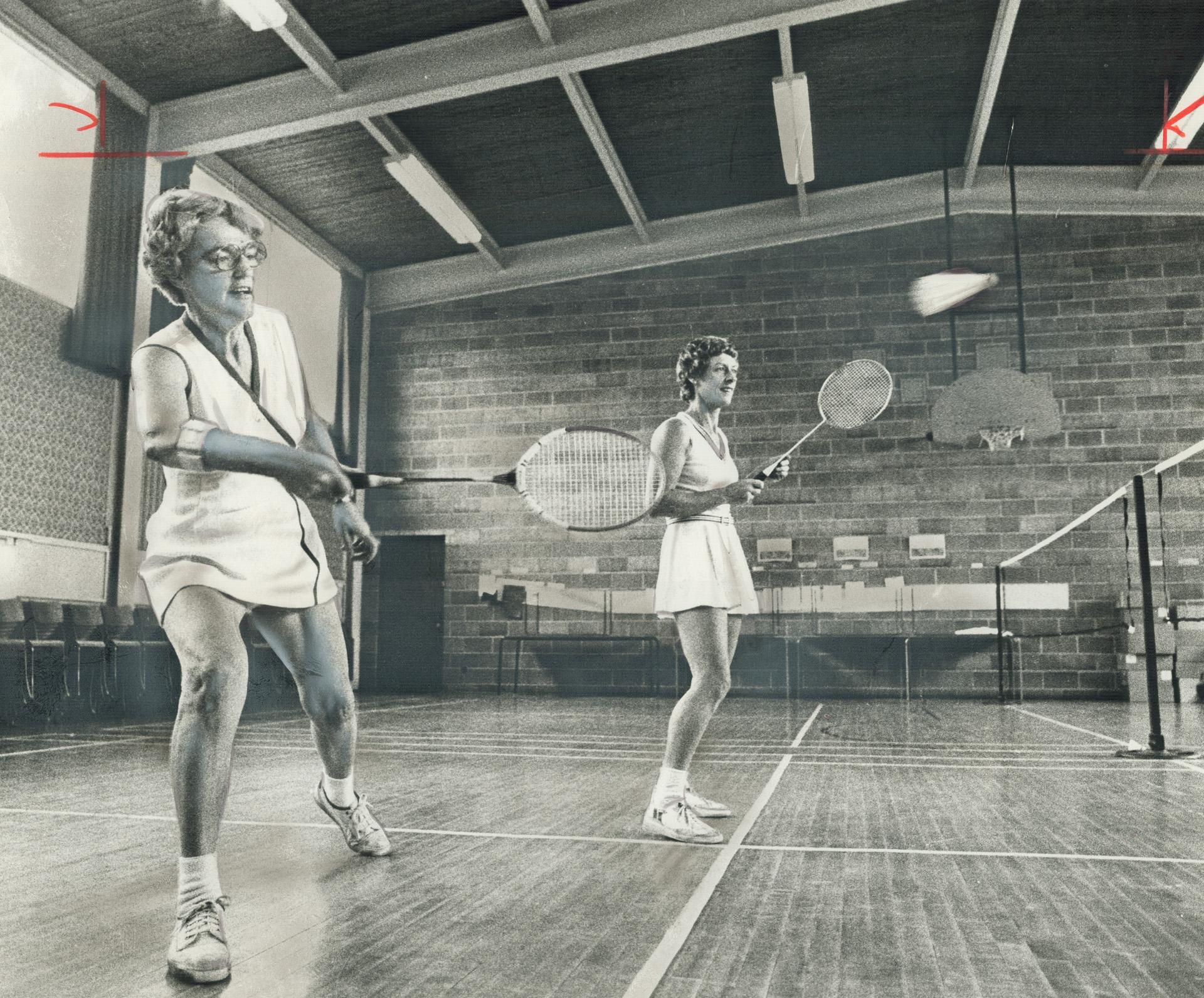 Two women playing badminton in a gymnasium hall.