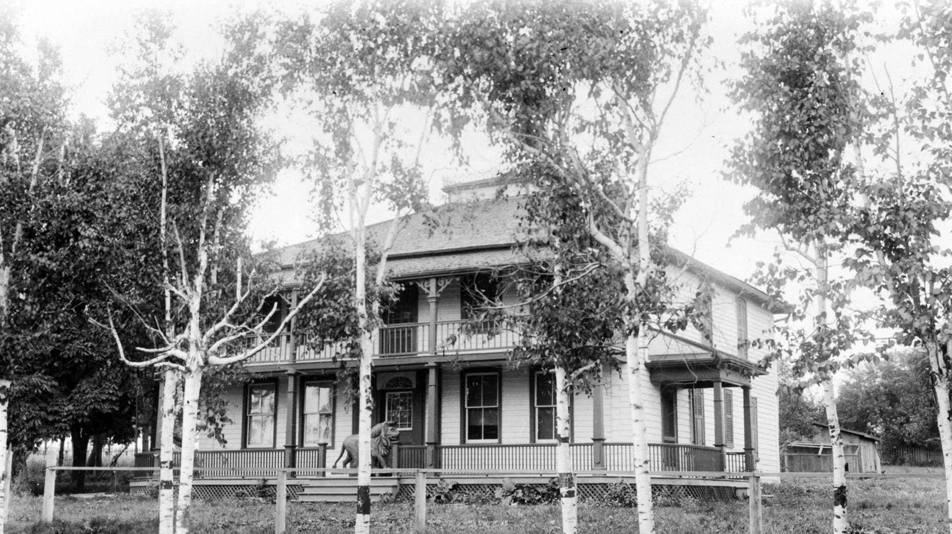 Black and white photo of a massive two-storey home with birch trees lining the property. Each storey of the house has a wrap-around balcony, and a statue of a lion sits at the entrance.
