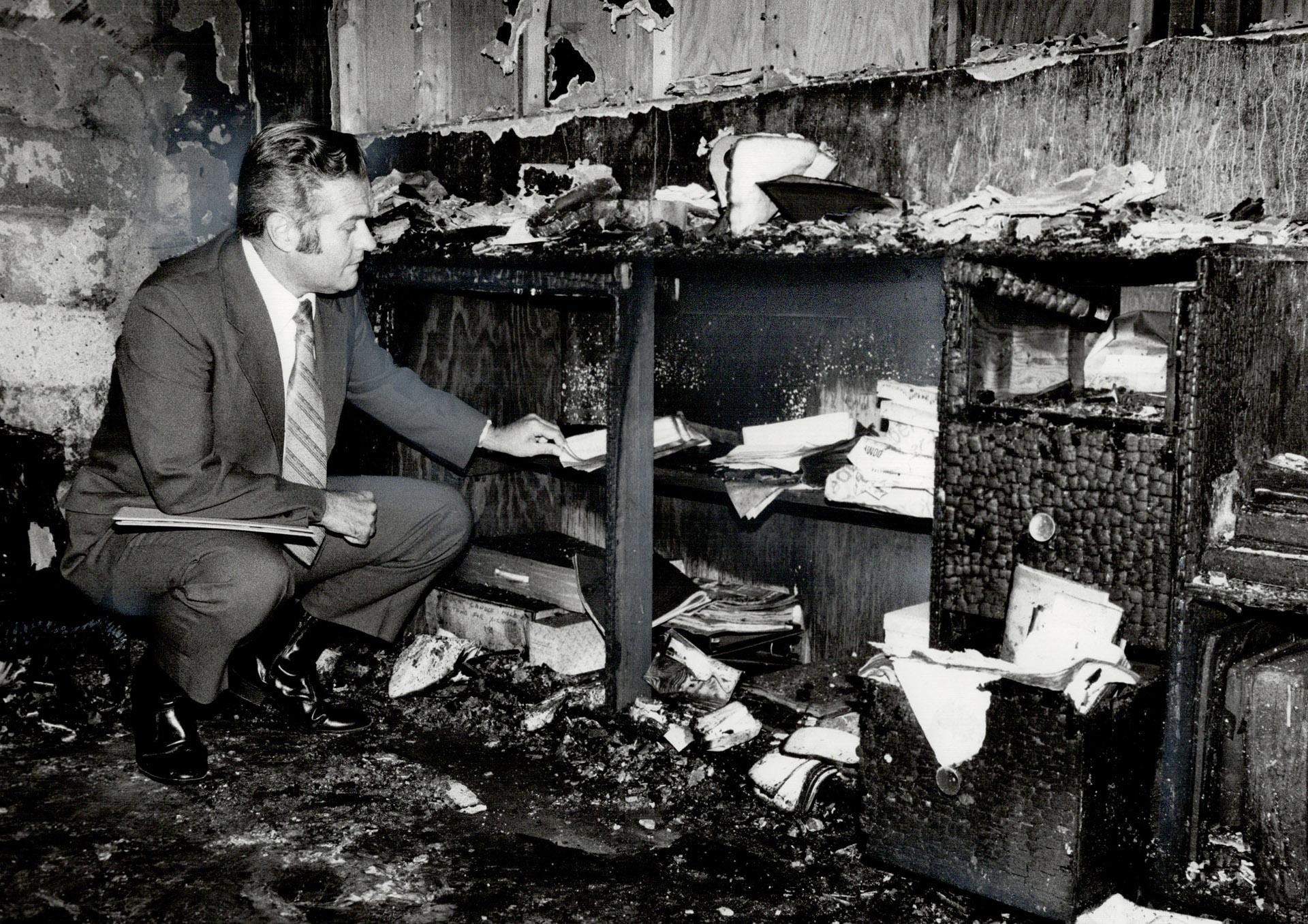 A suited man crouches, touching papers sitting inside of a cabinet, which is burnt along with much of its contents. Every surface is coated in ash and soot. 