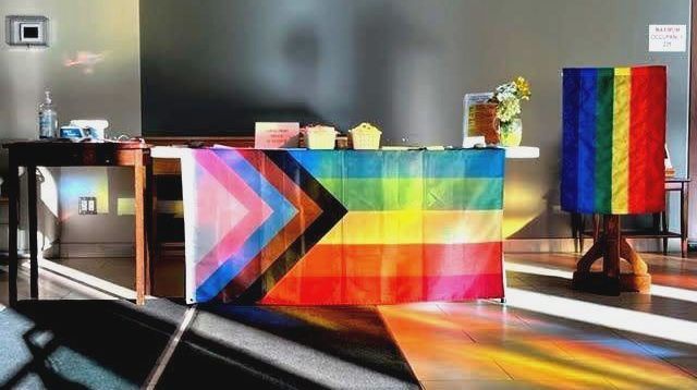 Lansing's welcome table, which holds face masks, flowers, and has a progress pride flag hung from it. Next to the table is another rainbow pride flag hanging from a lectern.