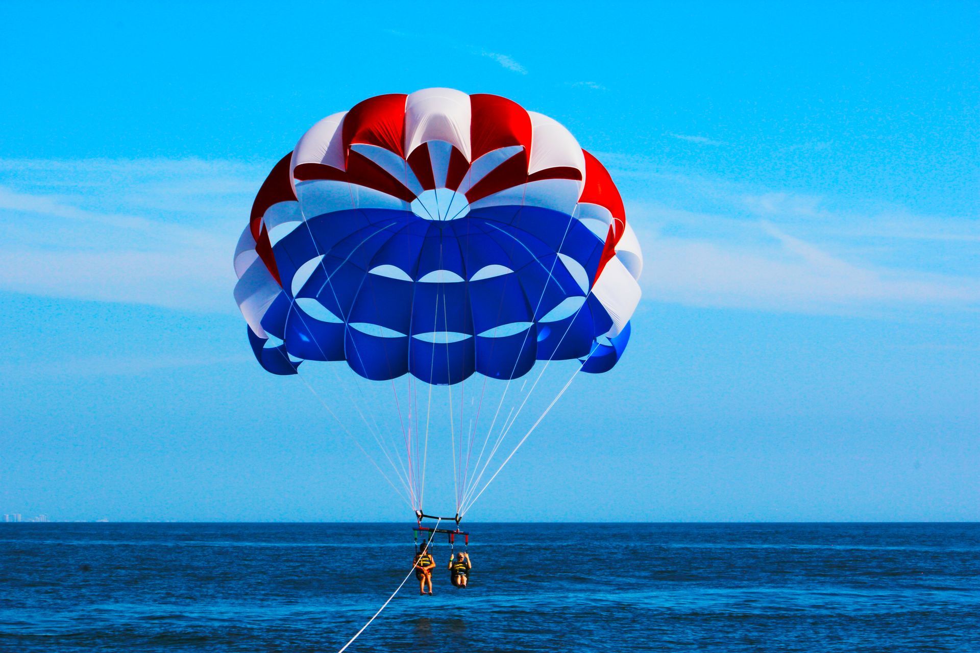 Two people parasailing over the ocean under a red, white, and blue parachute on a sunny day.
