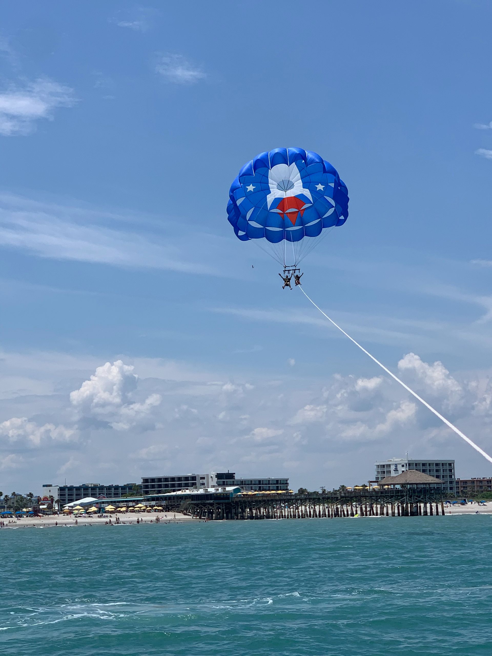 Blue parasail with two people over ocean, beach, and buildings.