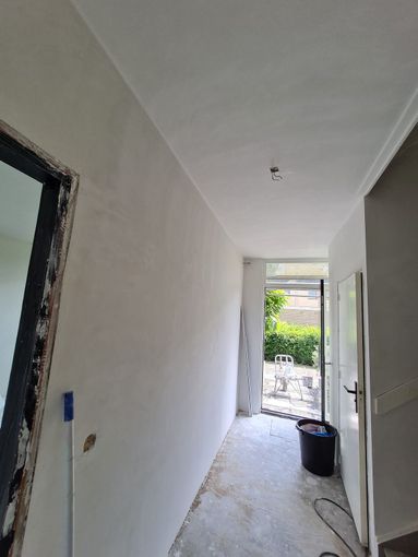 Smalle gang in renovatie met witte muren, onafgewerkte vloer en een open deur naar een lichte tuin.