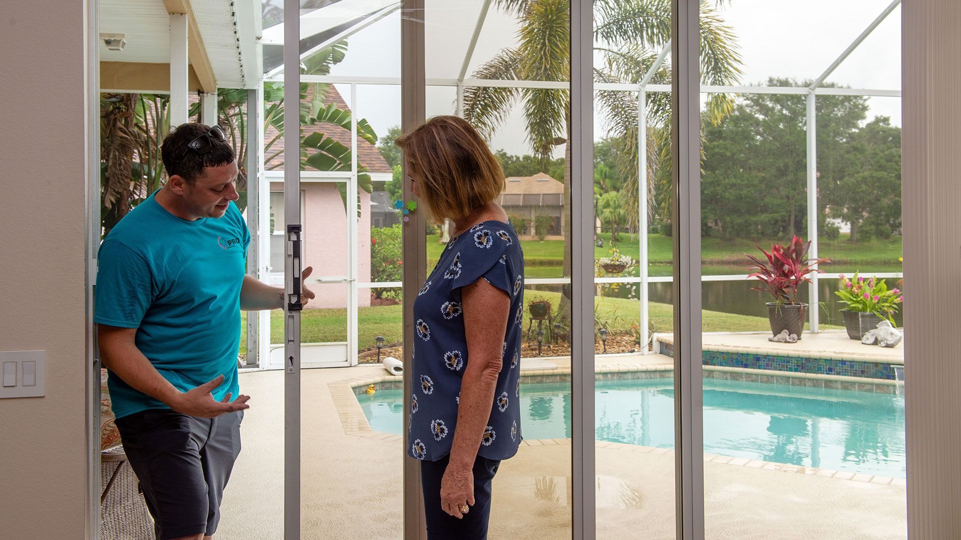A person in a blue shirt demonstrates a sliding glass door to another individual overlooking a backyard pool.