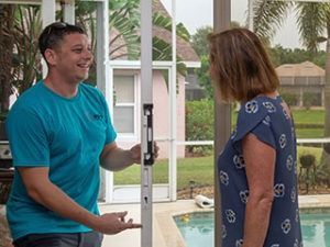 A person in a blue shirt holds a level against a door frame while standing outdoors by a pool with another person.