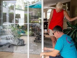 A person in a blue t-shirt kneeling to repair a white sliding glass door while another person observes from indoors.