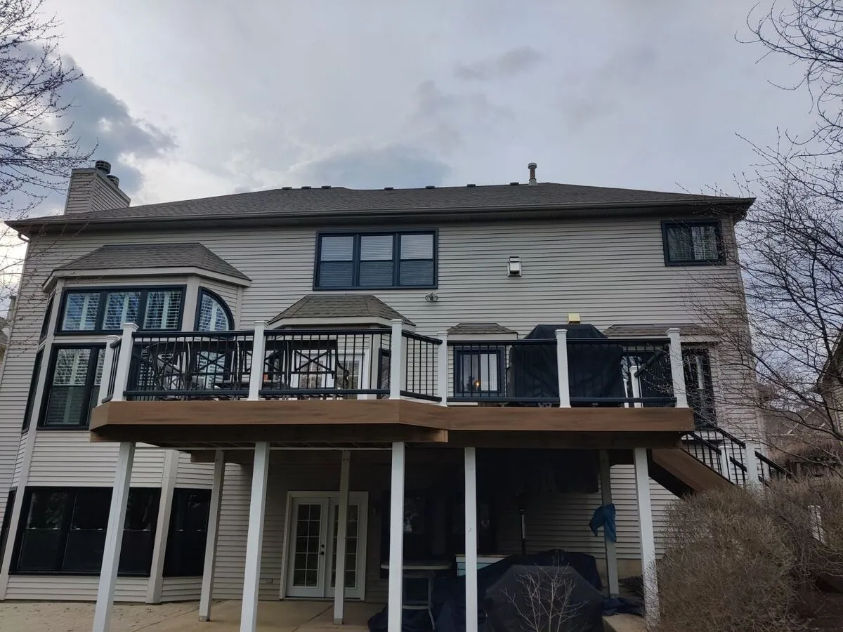 Back of a two-story beige house with a brown deck, black railing, and cloudy sky.