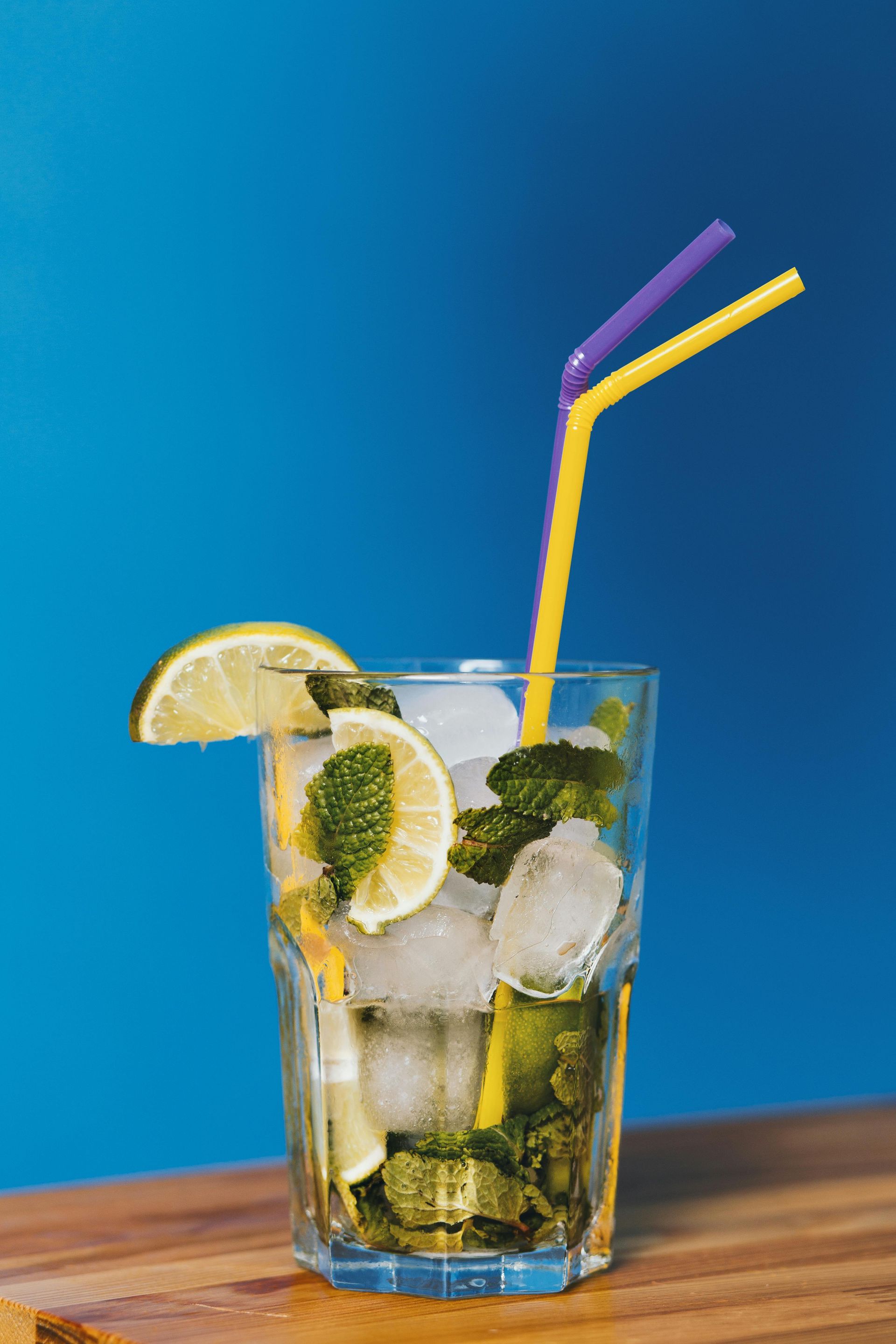 Mojito in a glass with mint, lime, ice, and straws, against a blue background.