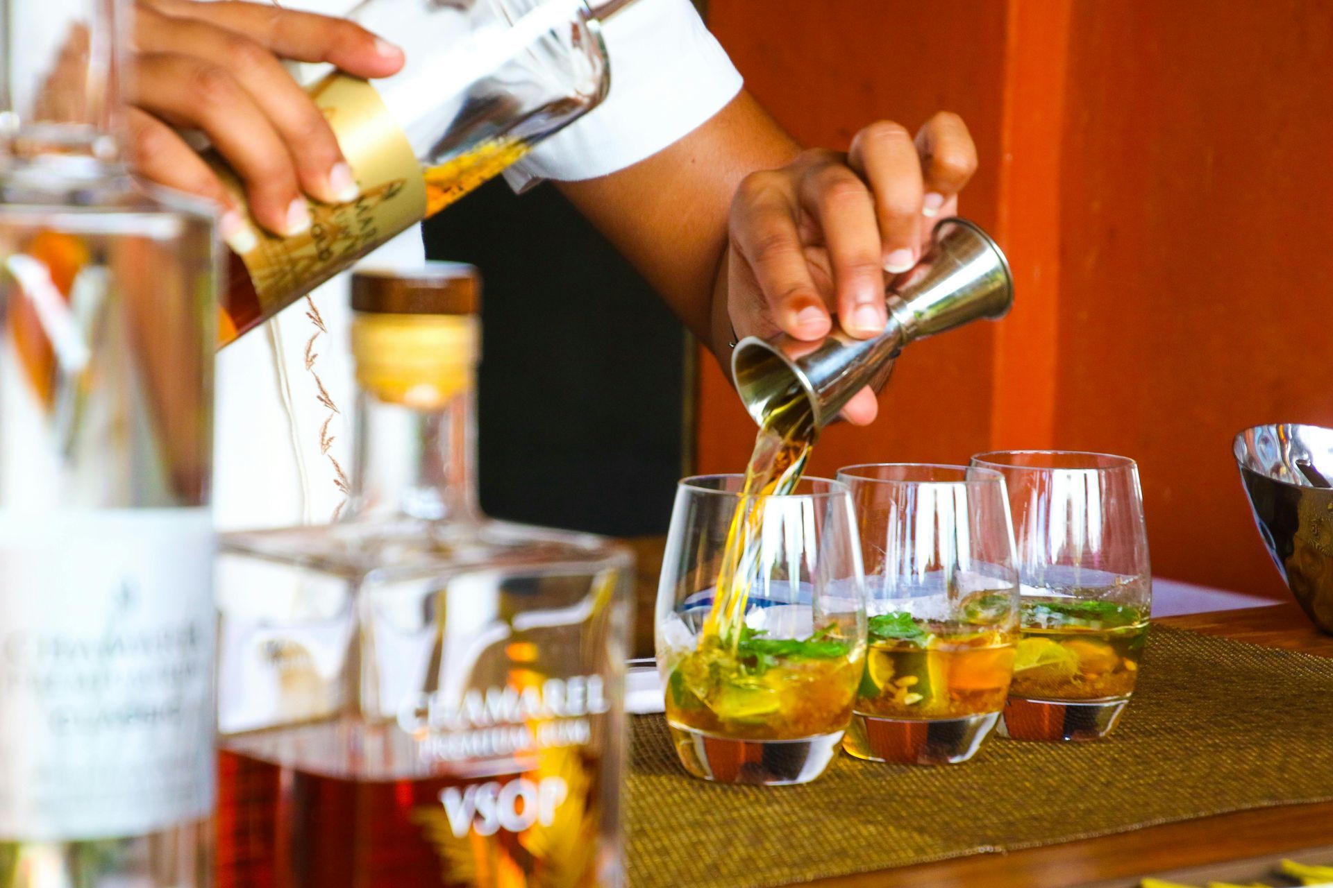 Bartender pouring amber liquid from a jigger into three glasses with mint and ice, behind liquor bottles.