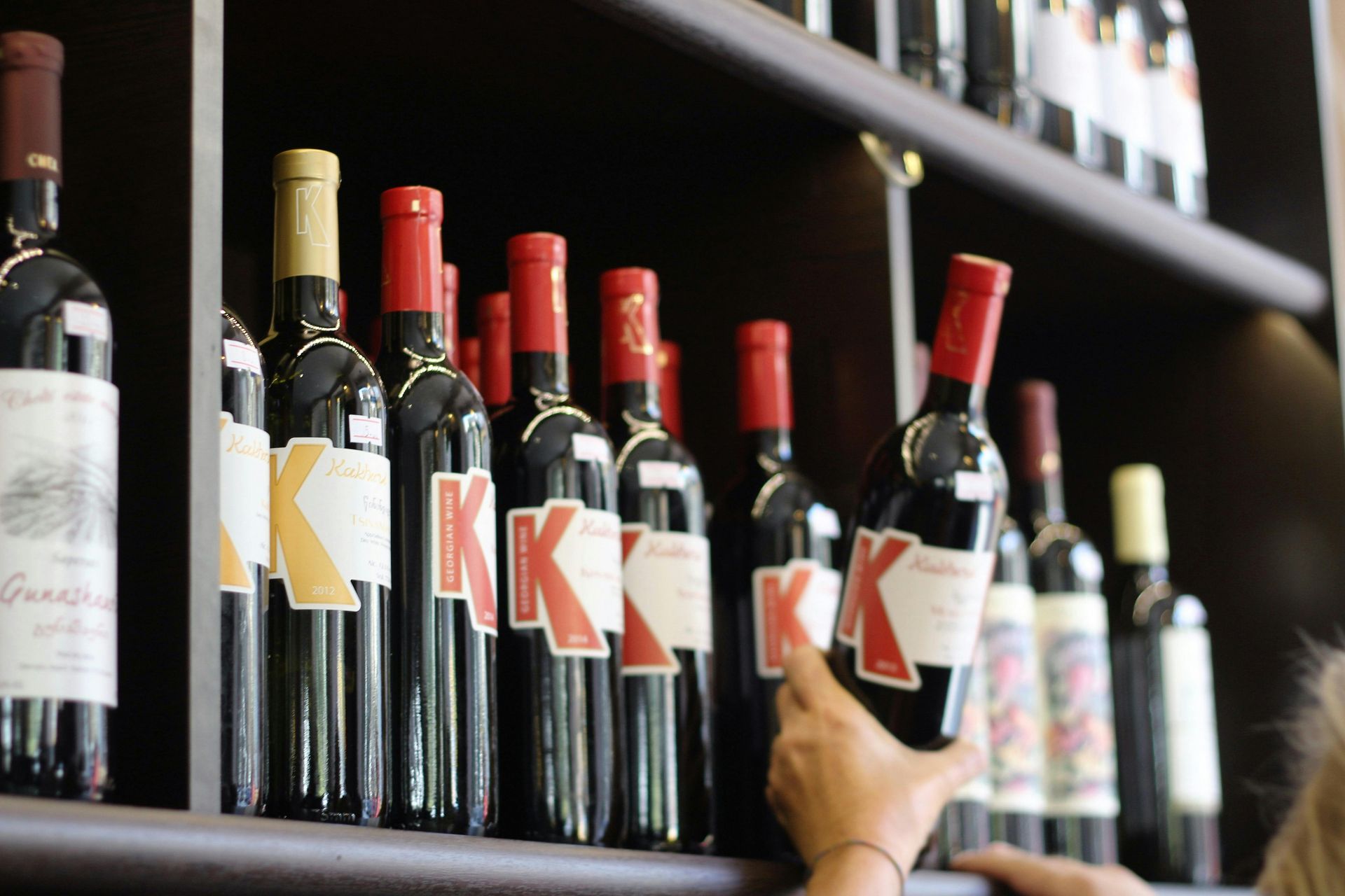 Person selecting a bottle of red wine from a shelf filled with wine bottles with red and gold labels.