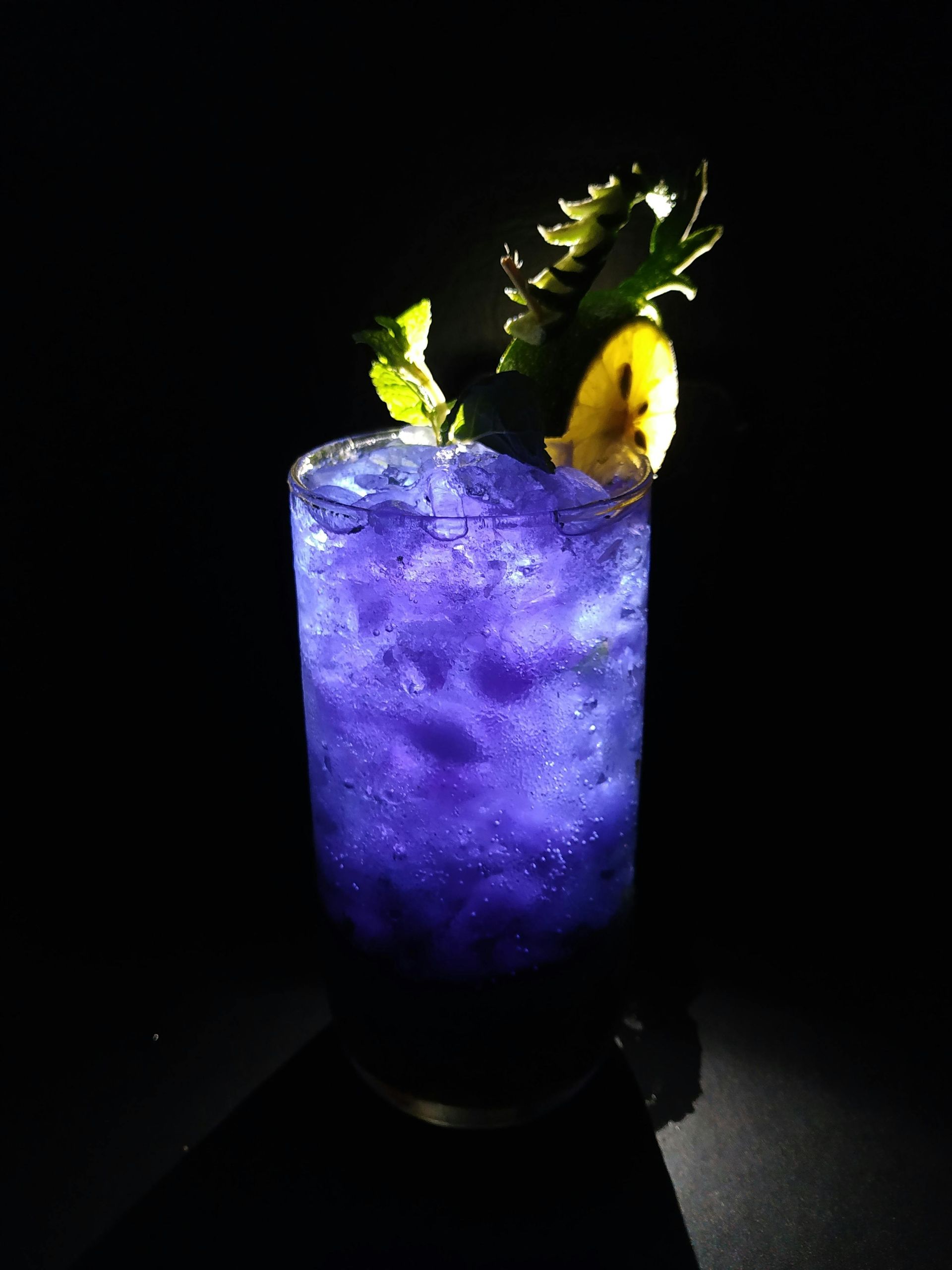 Purple cocktail in a tall glass, garnished with mint and a lemon peel against a black background.