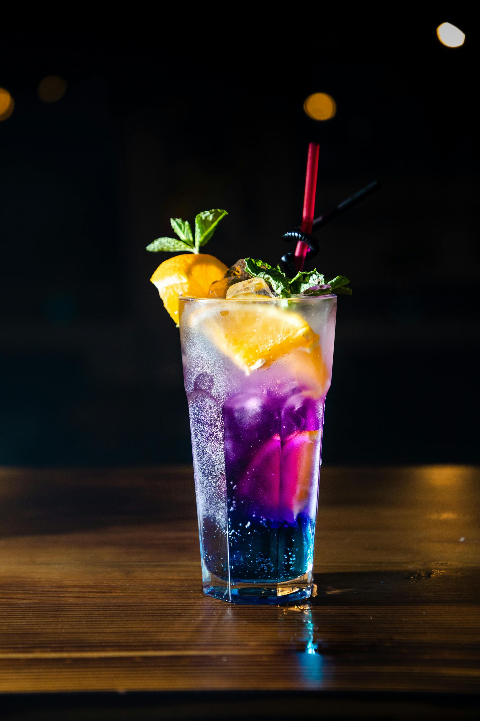 Colorful cocktail in a tall glass, garnished with orange slice and mint, set on a wooden surface against a dark background.