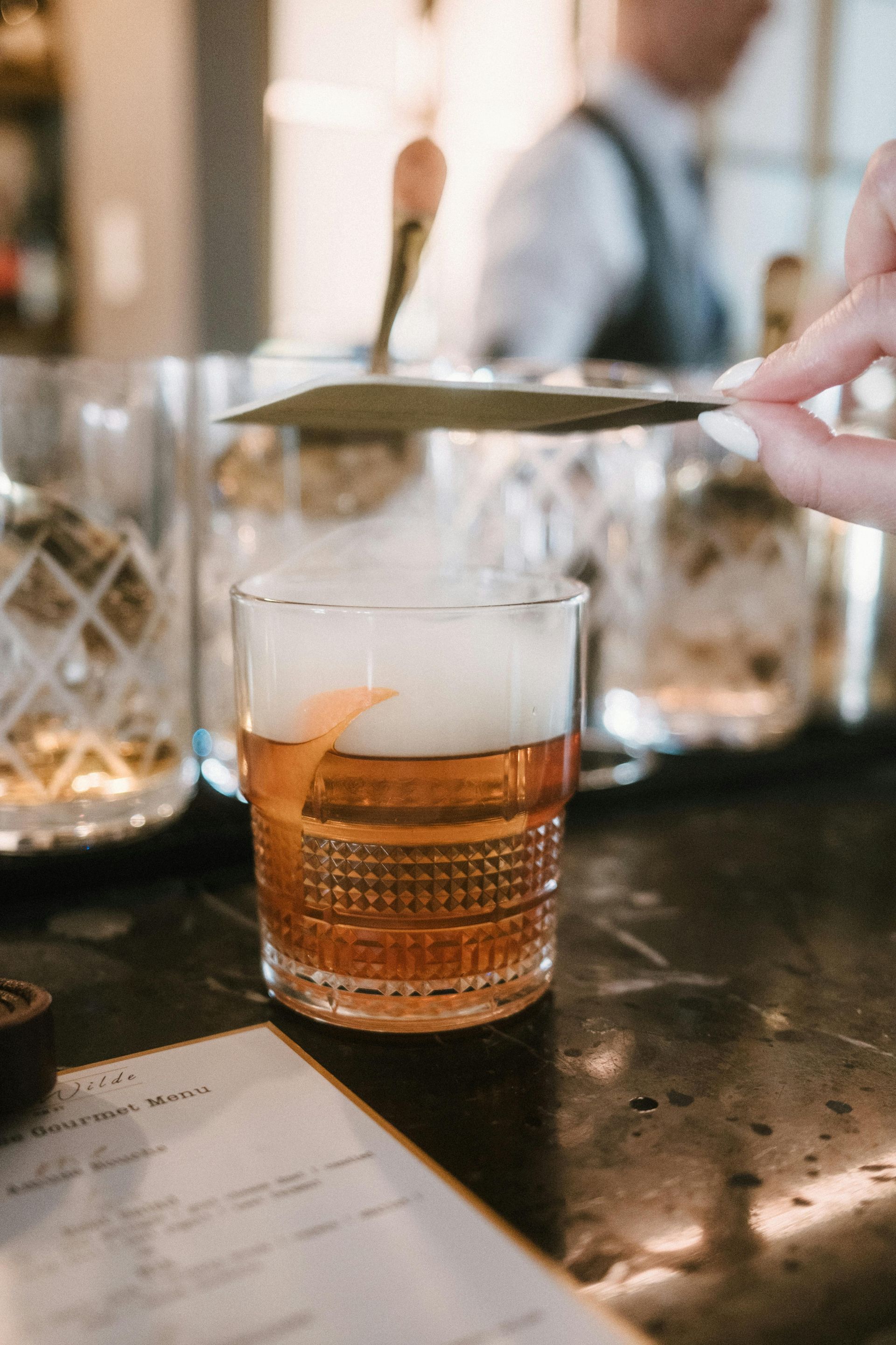 Cocktail being smoked with a hand holding a lid. Orange peel garnish. Bar setting.