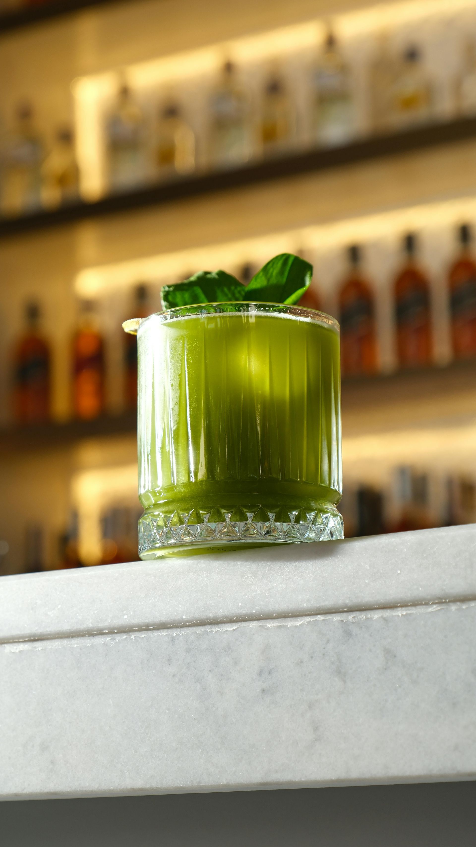 Green cocktail in a ribbed glass, garnished with basil, on a bar.