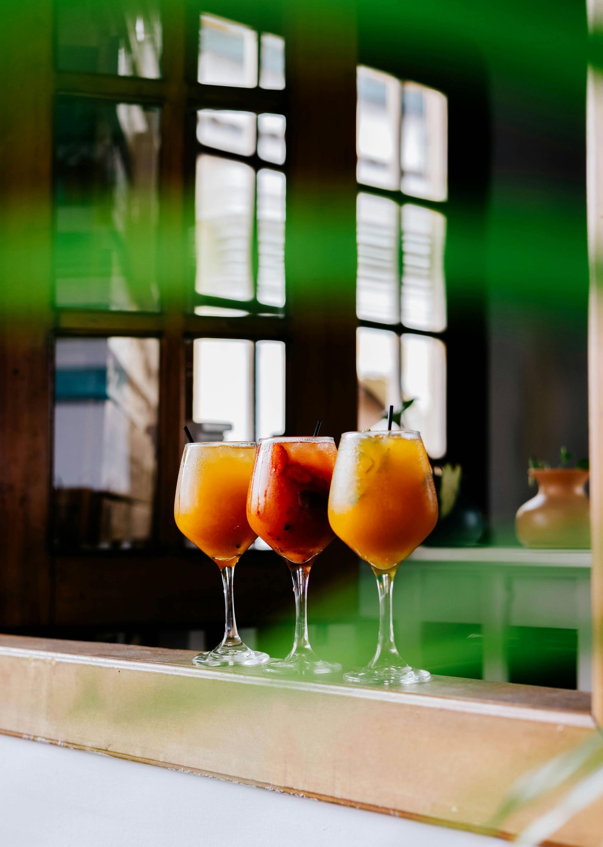 Three orange cocktails in stemmed glasses on a windowsill, with a window and greenery blurred in the background.