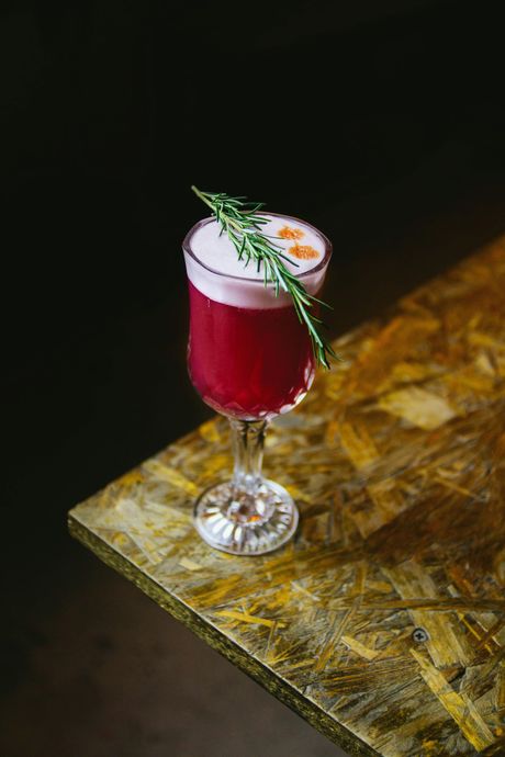Cocktail in a stemmed glass with a frothy top, garnished with rosemary sprig, on a textured wooden surface.