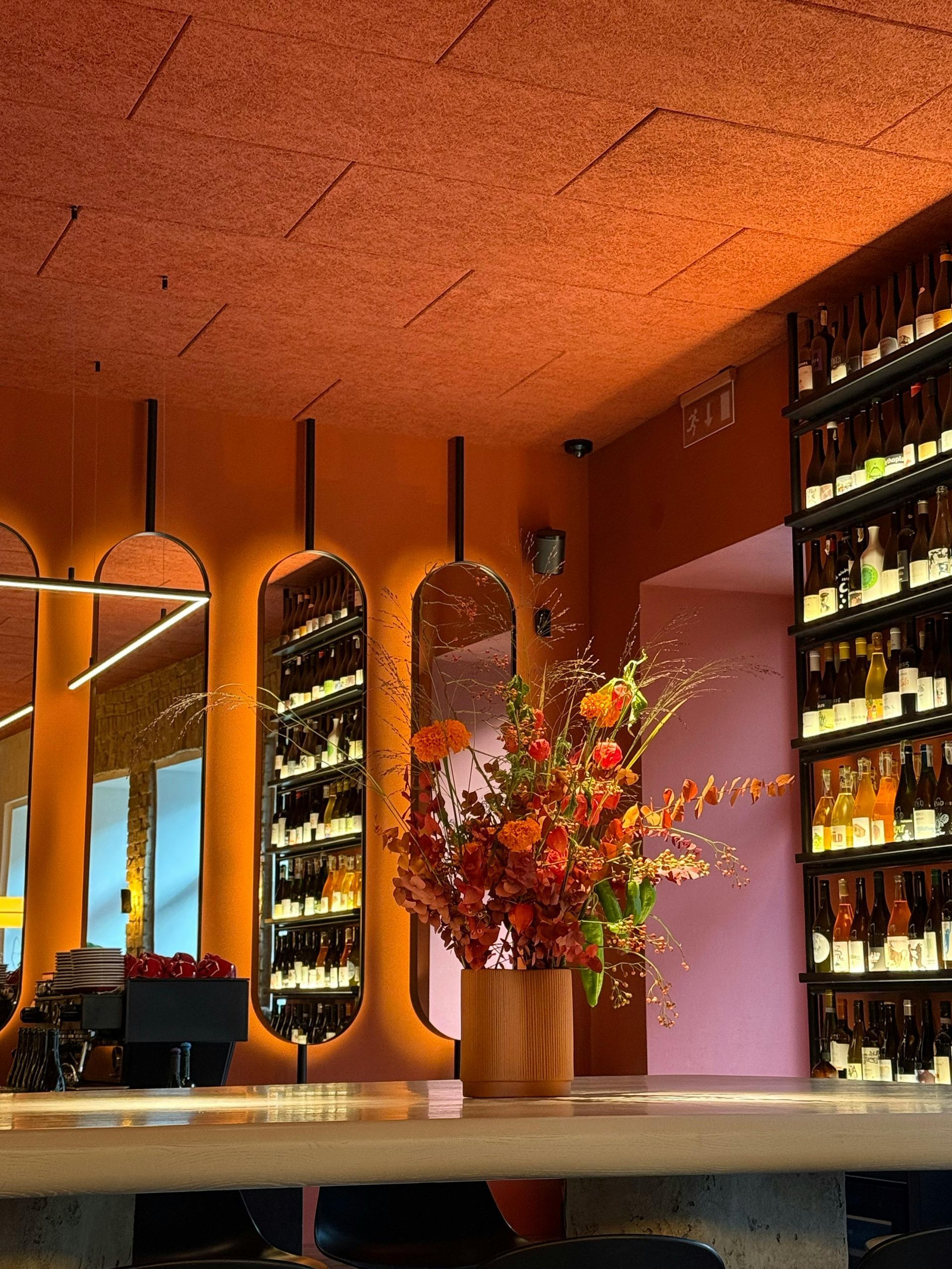 Bar interior with orange walls, mirrors, wine bottles, and flowers.