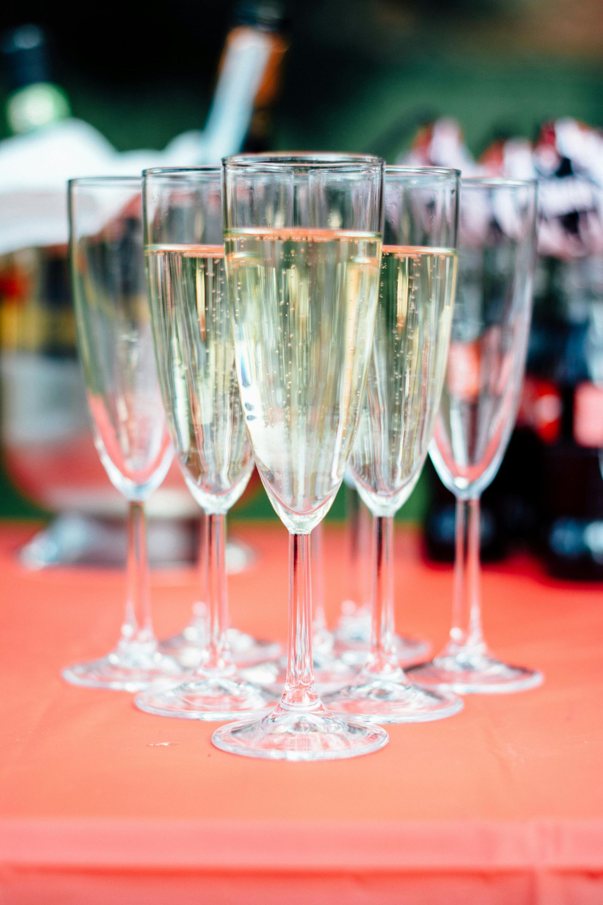 Champagne flutes filled with bubbly drink, on a red surface, other bottles in the blurry background.