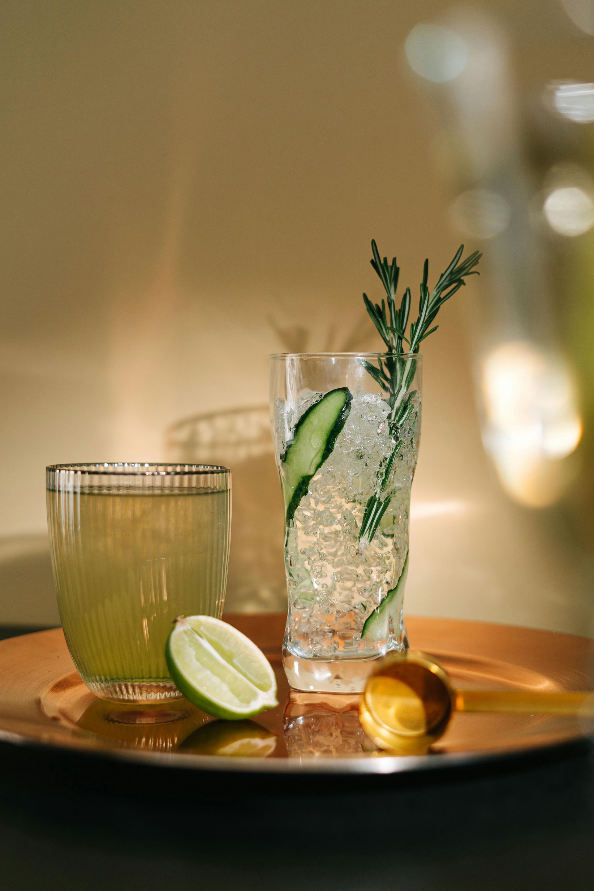 Two cocktails on a copper tray. One tall, garnished with cucumber and rosemary, one short, yellow liquid. Lime slice.