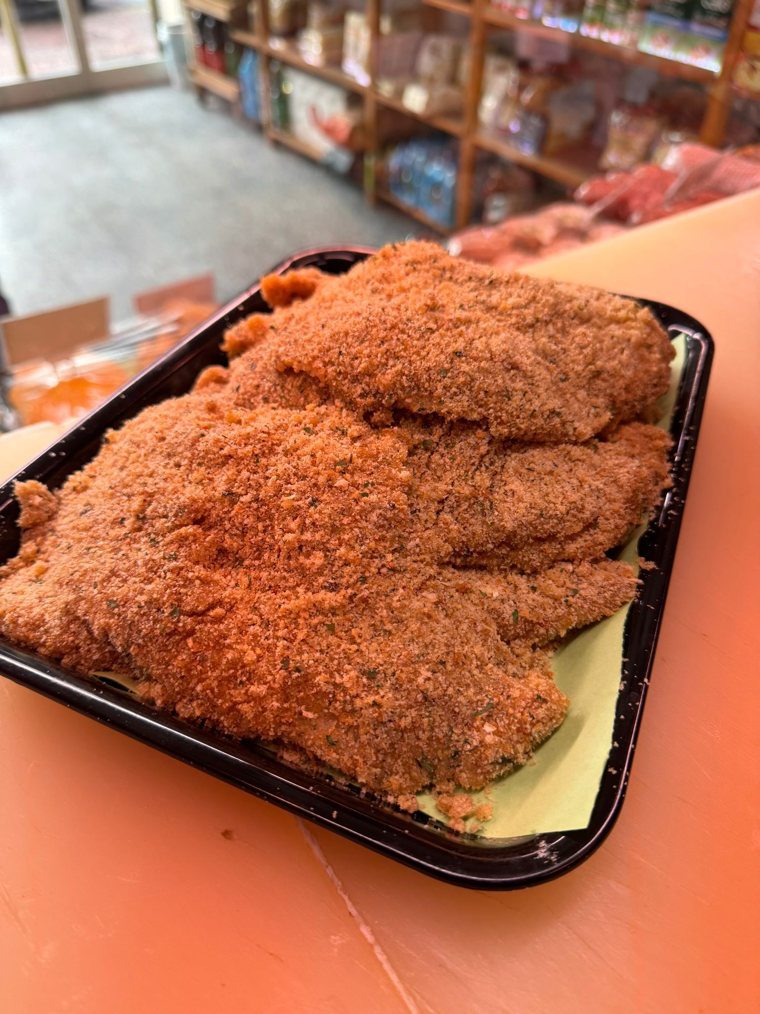 Filetti di pesce impanati su un vassoio nero, appoggiati su carta gialla, in un ambiente da negozio.