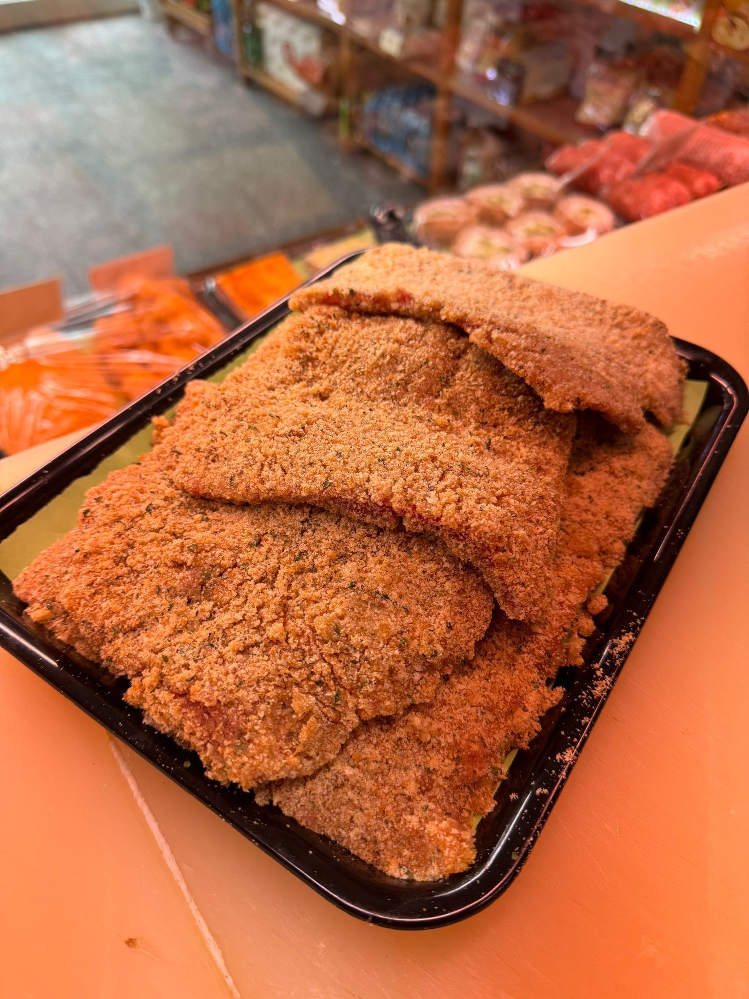 Cotolette impanate su un vassoio nero su un bancone, possibilmente in una macelleria.
