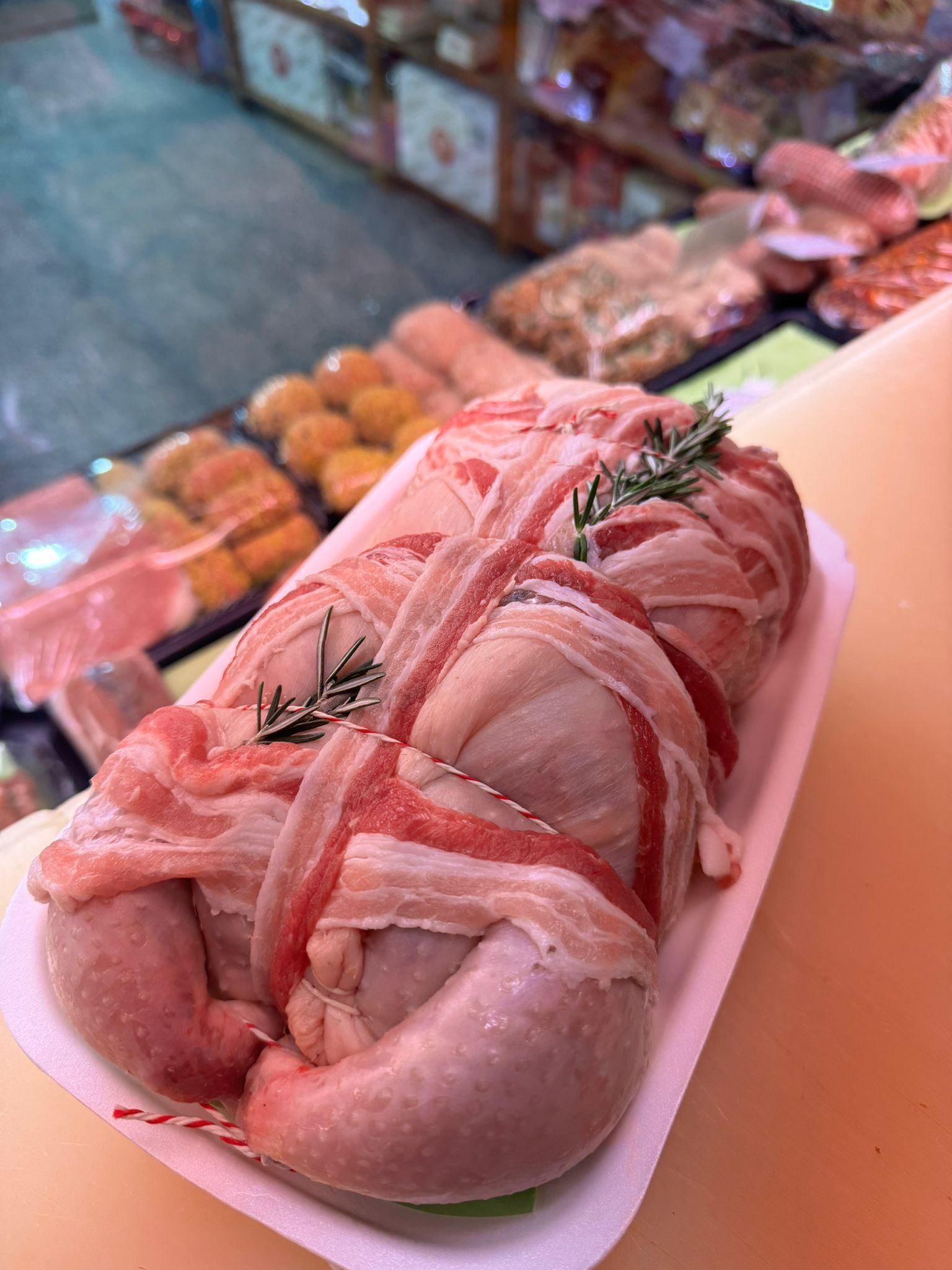 Polpettone crudo avvolto nella pancetta, legato con spago e guarnito con rosmarino, su un vassoio bianco, in un banco gastronomia.