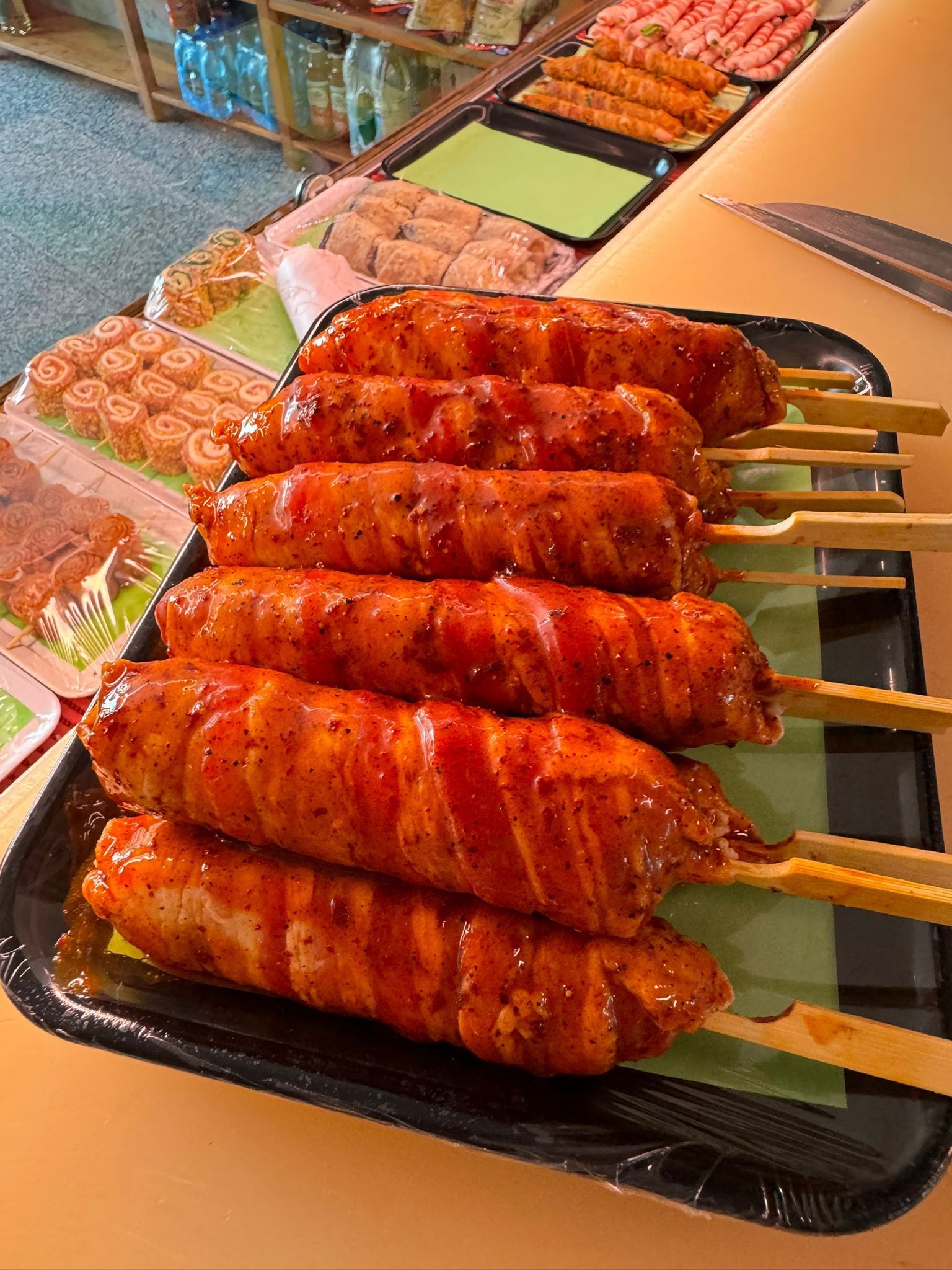 Spiedini di salsiccia grigliati, luccicanti di salsa, su un vassoio nero. Altri alimenti visibili sullo sfondo.