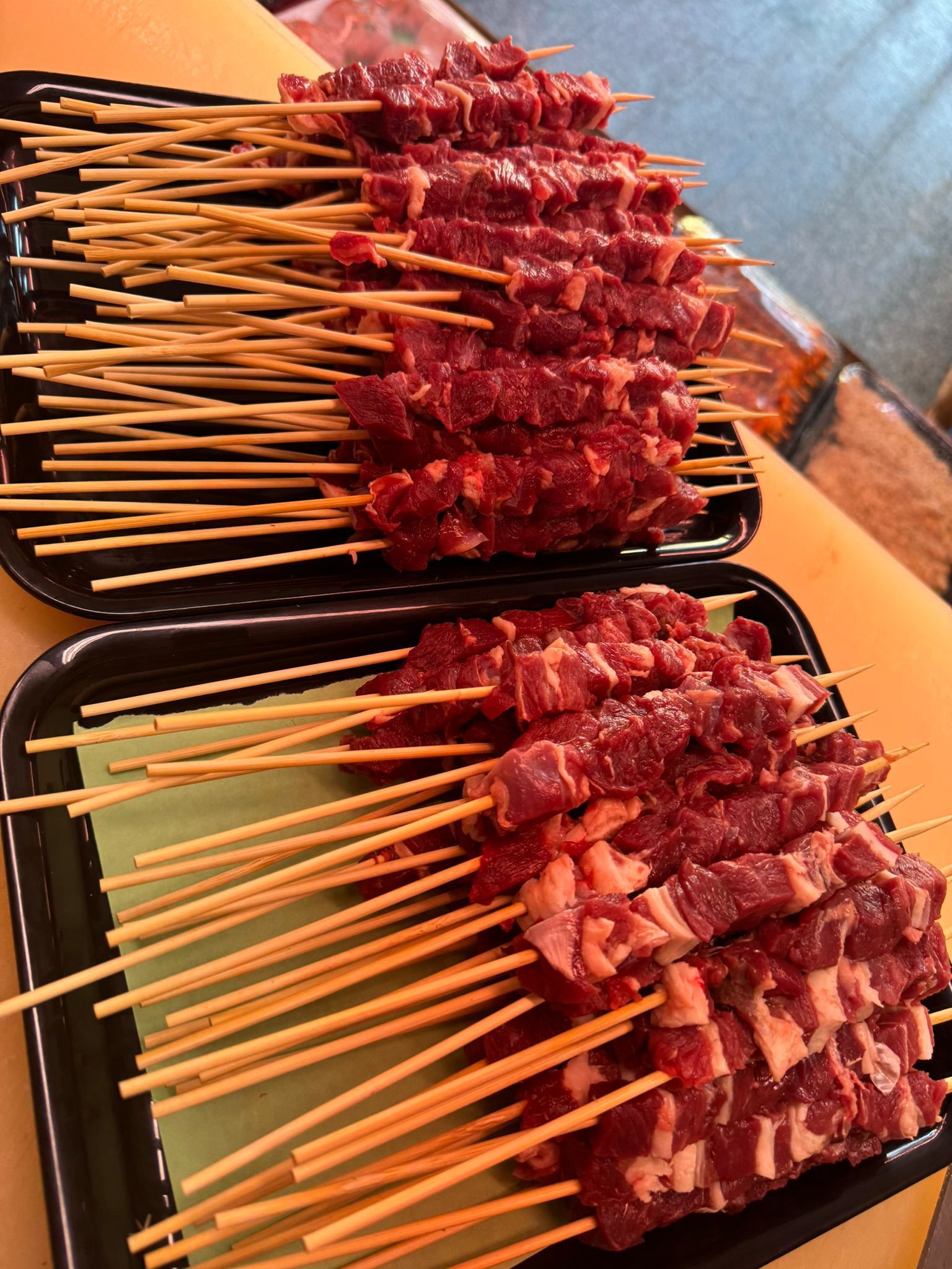 Spiedini di carne cruda in vassoi neri, pronti per essere grigliati.