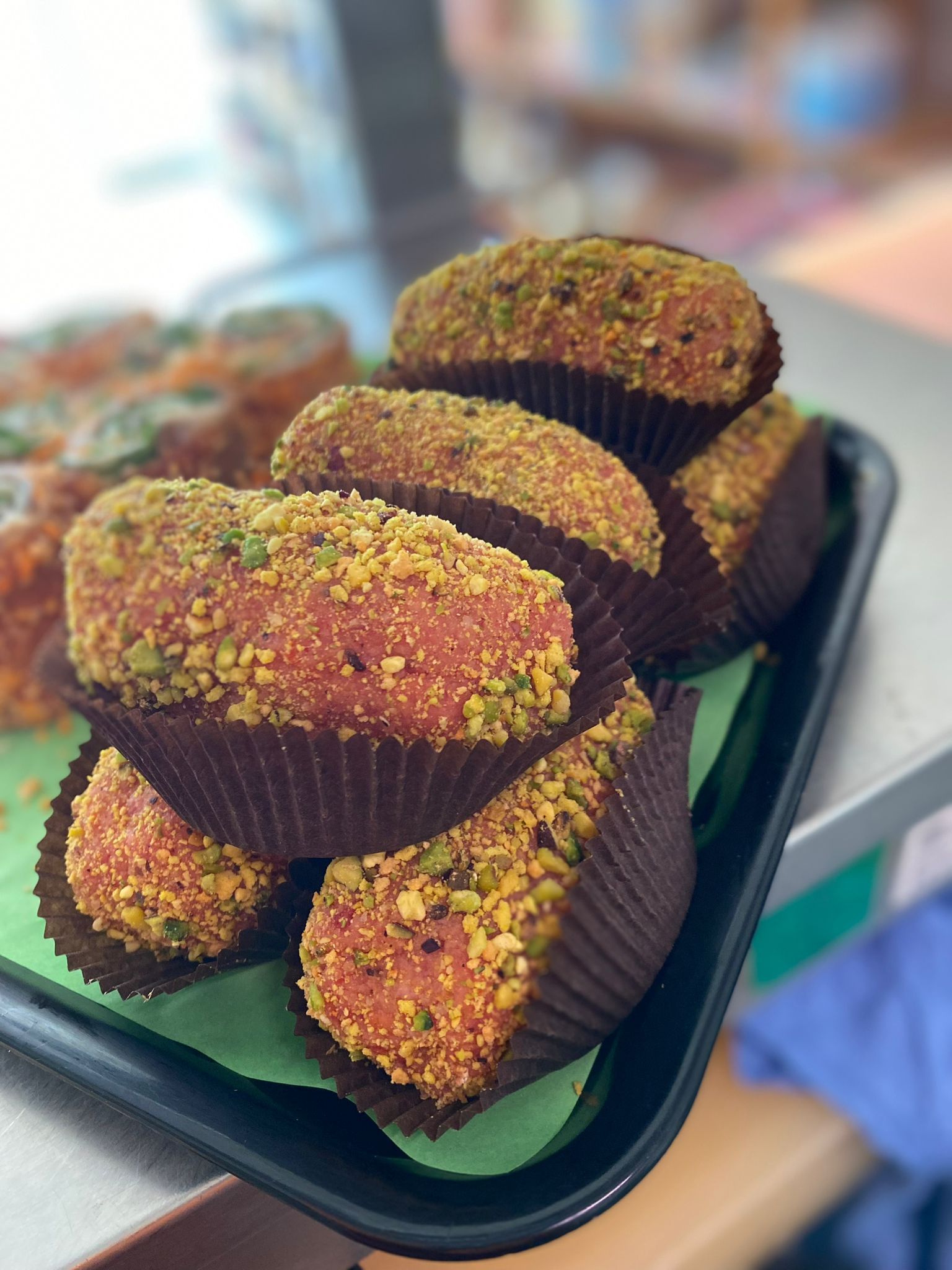 Vassoio di prodotti da forno: pasticcini rotondi e dorati ricoperti di briciole di pistacchio, in pirottini di carta.